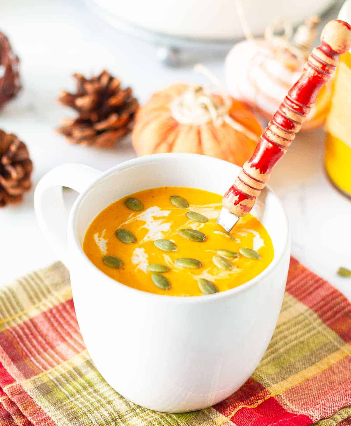 Roasted pumpkin soup in a white mug with a red spoon and ingredients in the background.