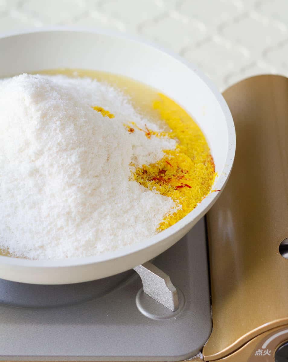 Adding desiccated coconut and saffron milk to the pan with sugar and khoya