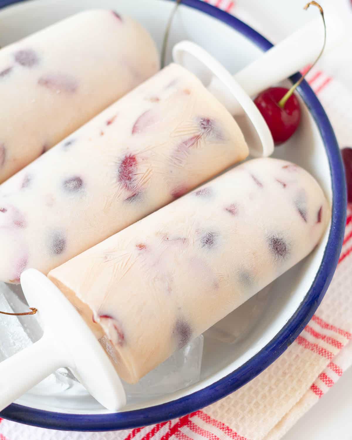 3 cherry yogurt popsicles in a dish with ice and cherries.