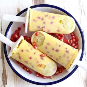 3 mango popsicles in a white bowl with pomegranates and ice.
