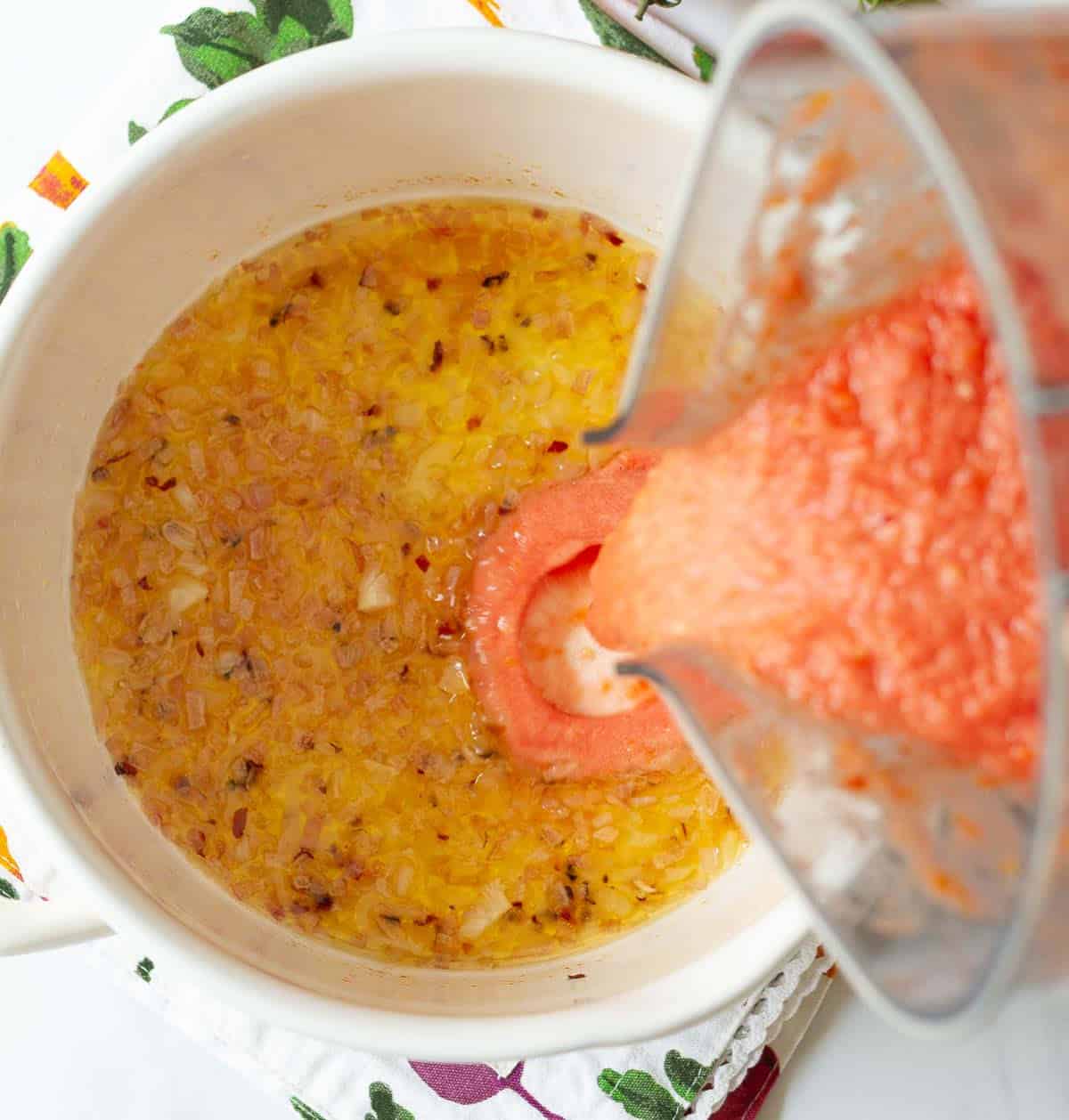 Tomato puree getting poured into a pot with sauteed onions from a blender jar.