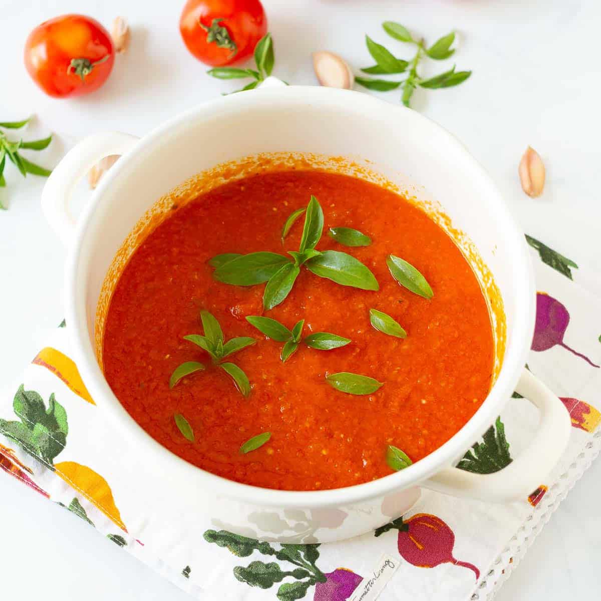 A pot with tomato sauce topped with fresh basil leaves and ingredients in the background.