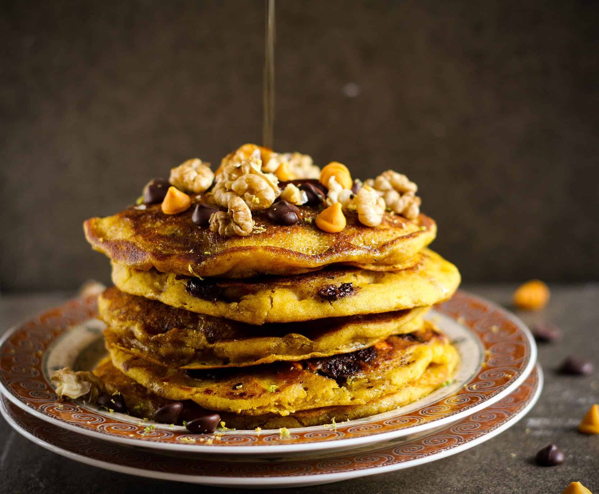 A stack of pumpkin pancakes with walnuts and chocolate chips getting drizzled with maple syrup.