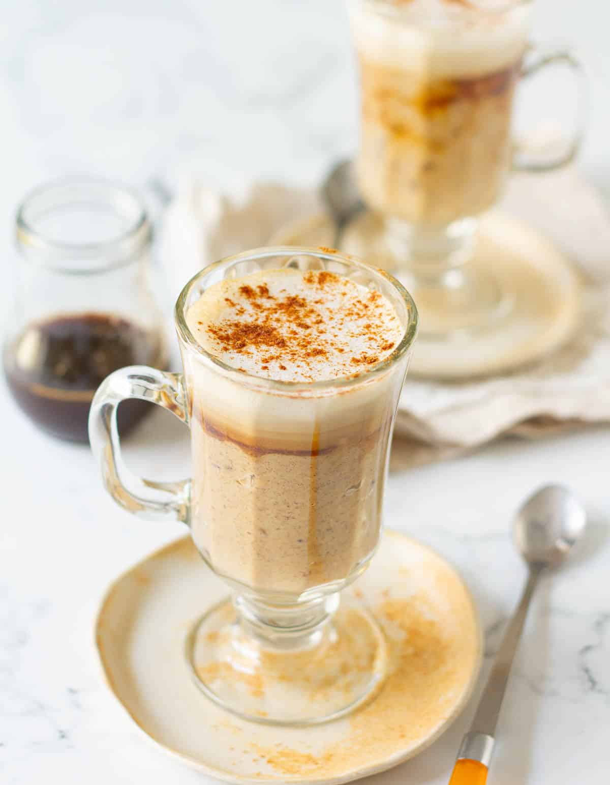 oatmeal latte in 2 glass mugs with brewed coffee and spoons in the background.
