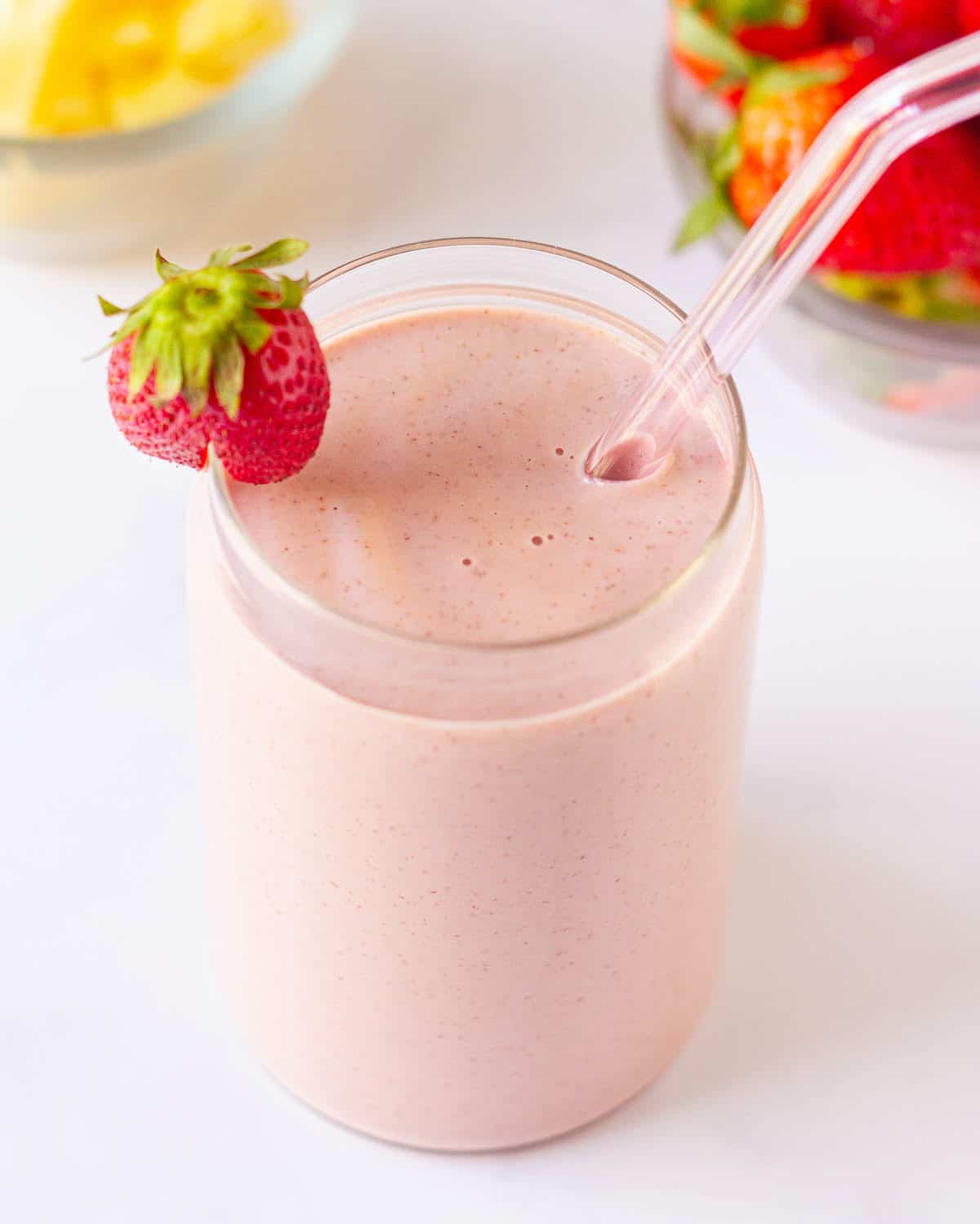 strawberry pineapple smoothie in a glass with a glass straw and strawberry on the rim.