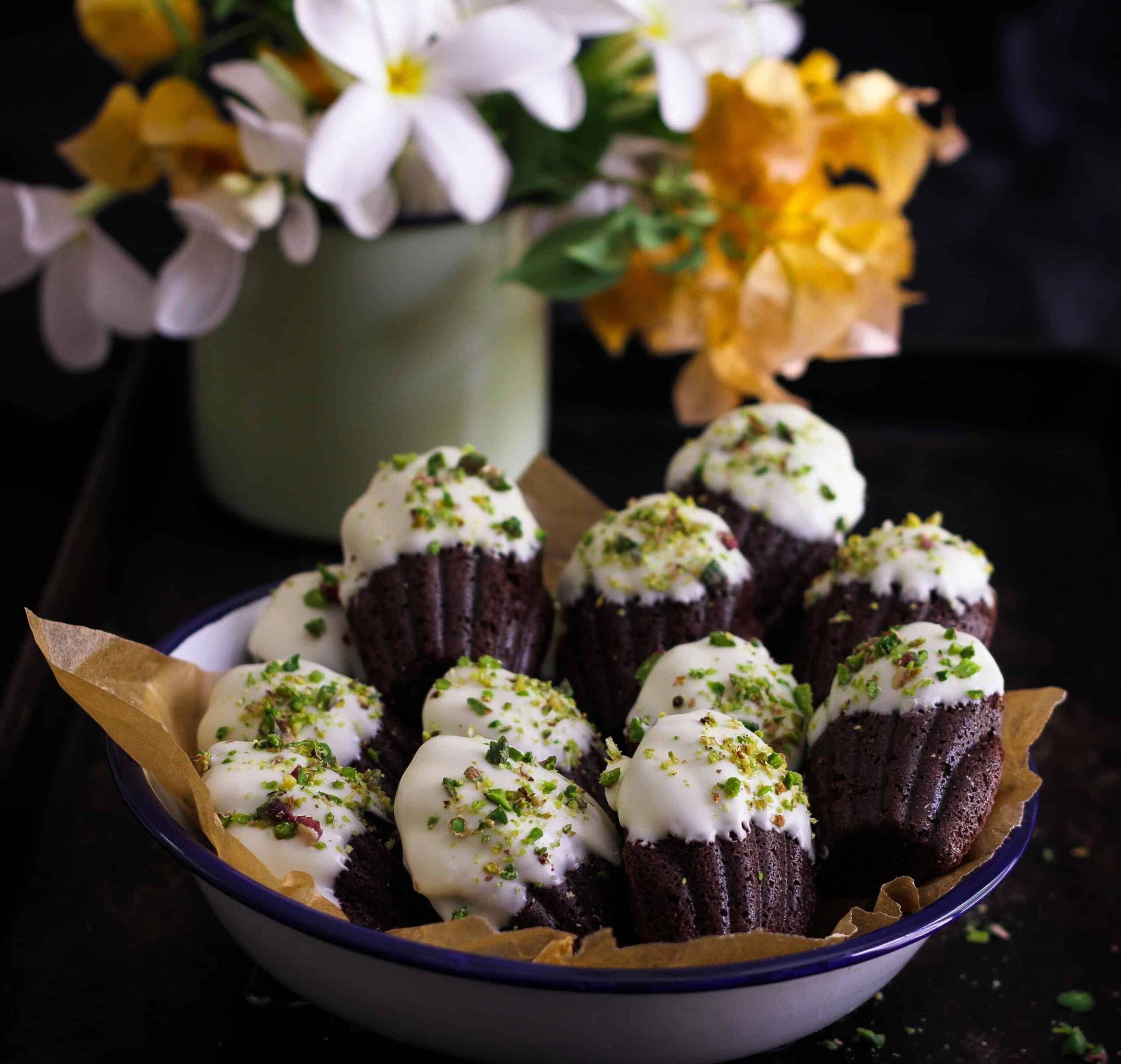 chocolate madeleines in a paper-lined bowl with flowers in the background.