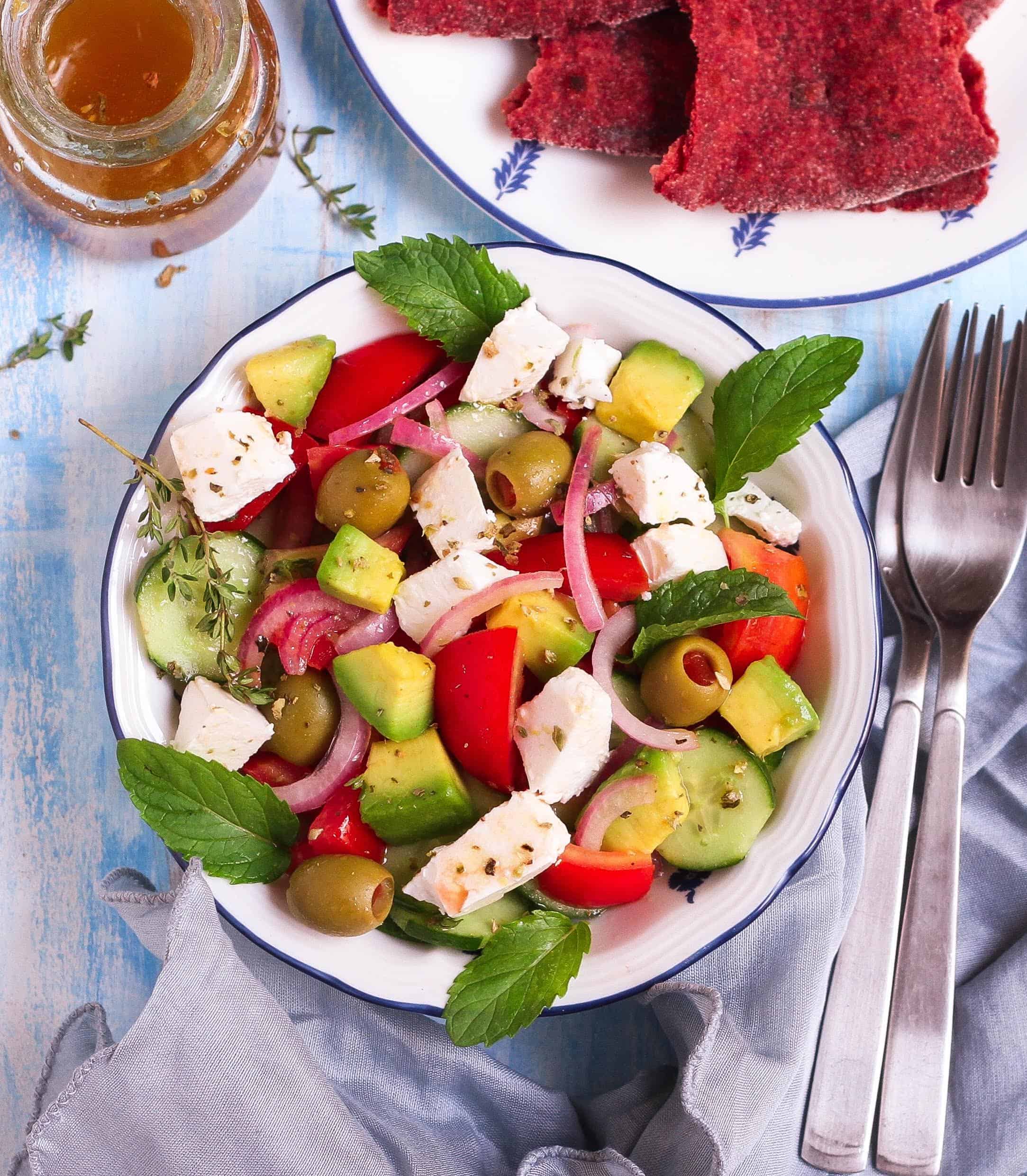 Greek salad with avocado in a bowl with the dressing and flatbread on the side.