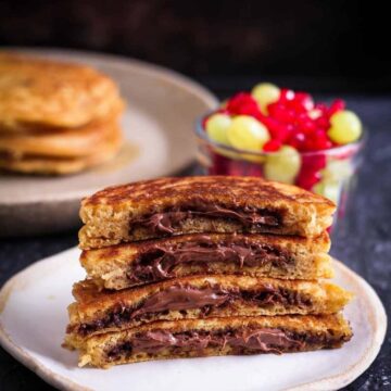A stack of half chocolate stuffed pancakes on a plate with fresh fruits around.