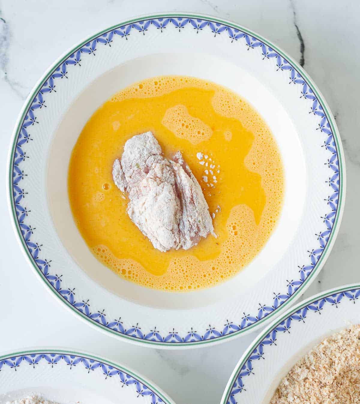 A flour coated chicken piece in a bowl of beaten eggs.