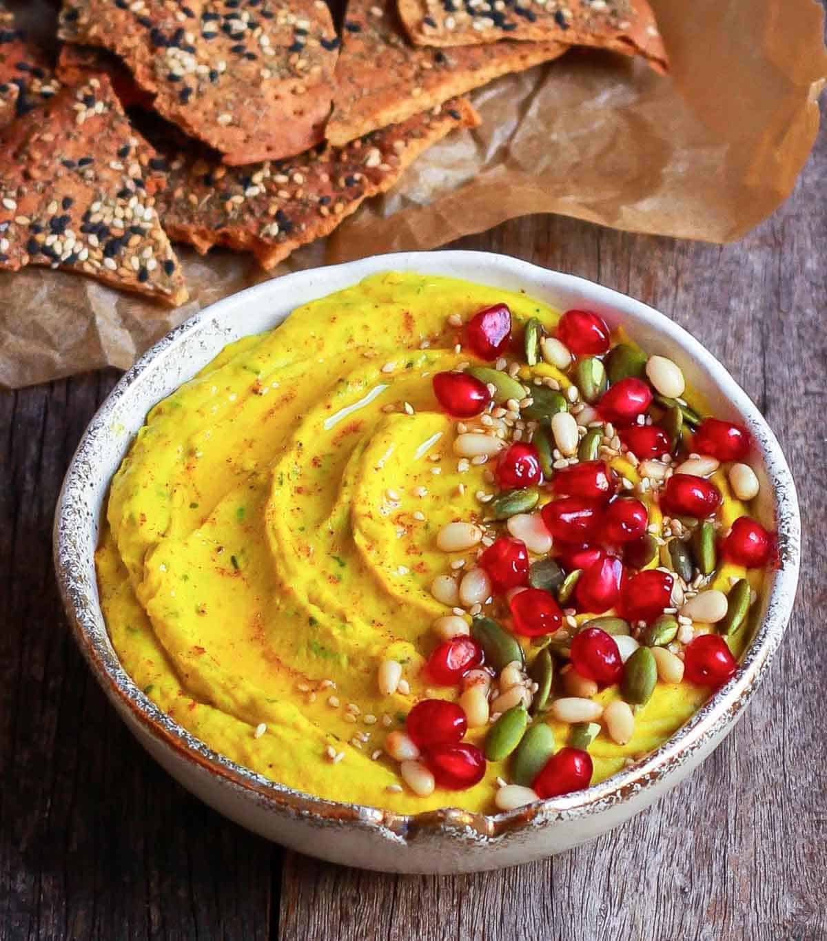 Hummus topped with nuts, pomegranates and pumpkin seeds in a bowl with crackers.