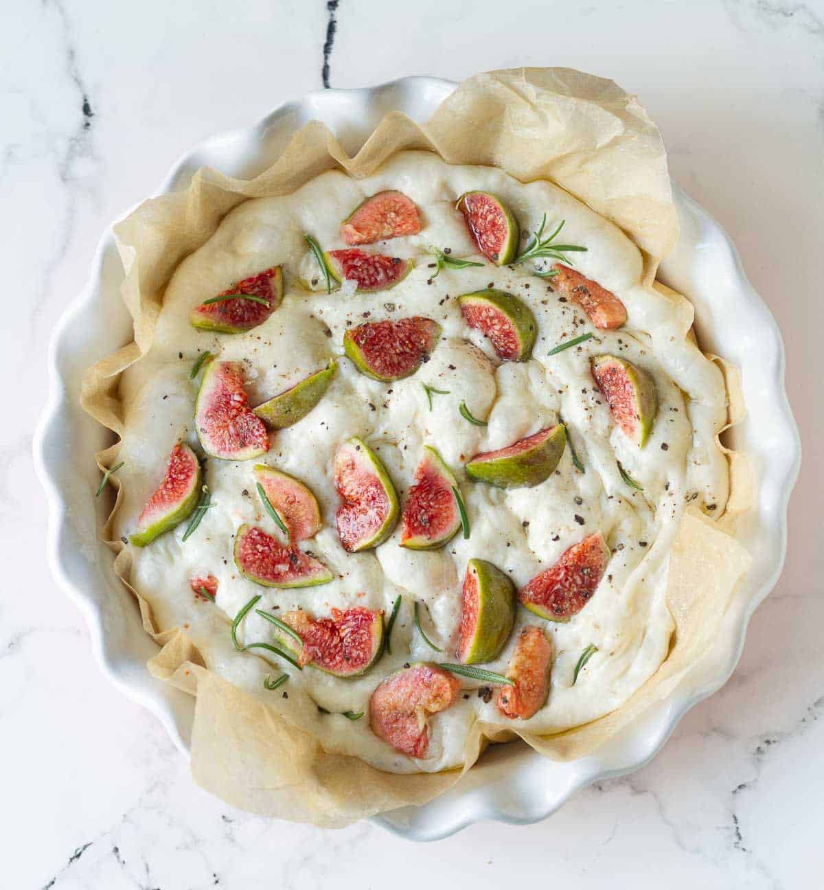 Dough topped with fresh figs, rosemary, salt, and pepper in a paper-lined dish.