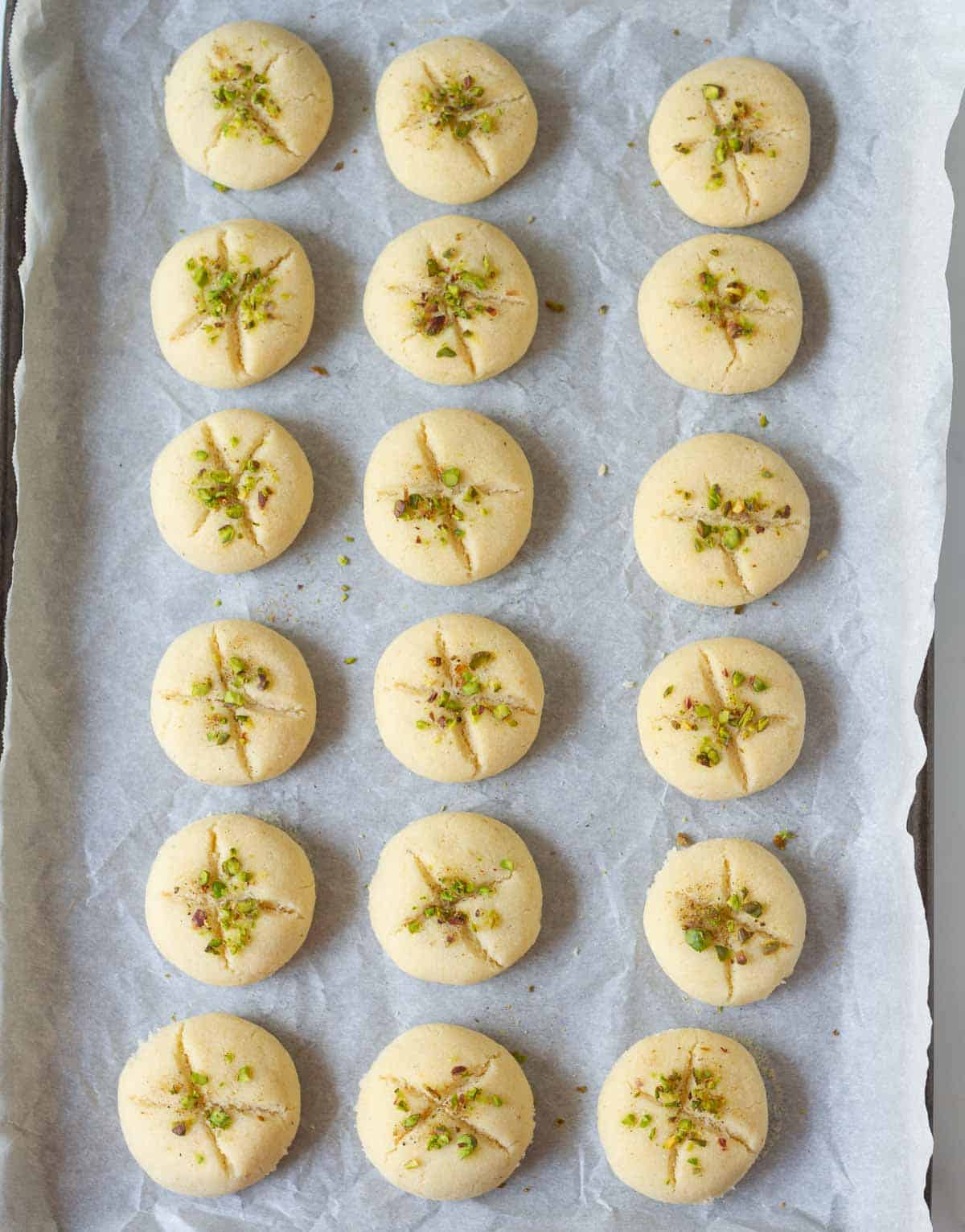 Baked cookies with pistachio and cardamom powder on top on baking paper lined tray.