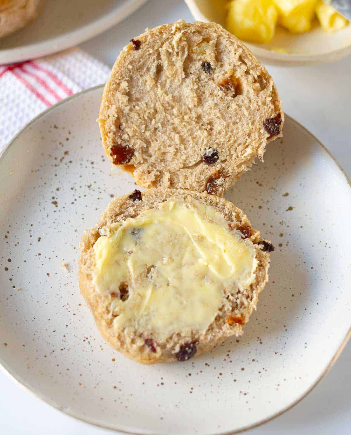 One hot cross bun halved on a plate with butter on one half.