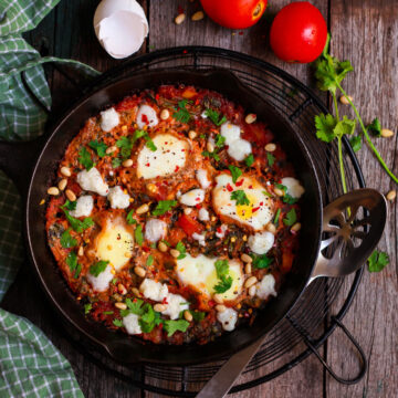 A cast iron pan with tomato sauce and cooked eggs with ingredients around.