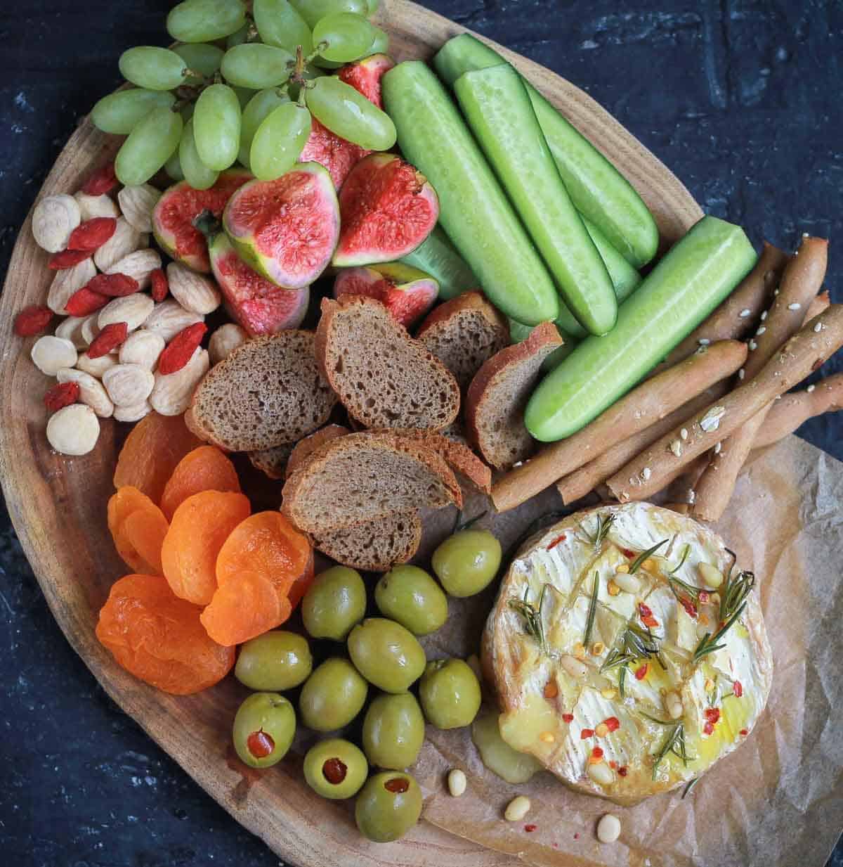 baked camembert on a wooden board with olives, crackers, veggies, nuts, and fruits.