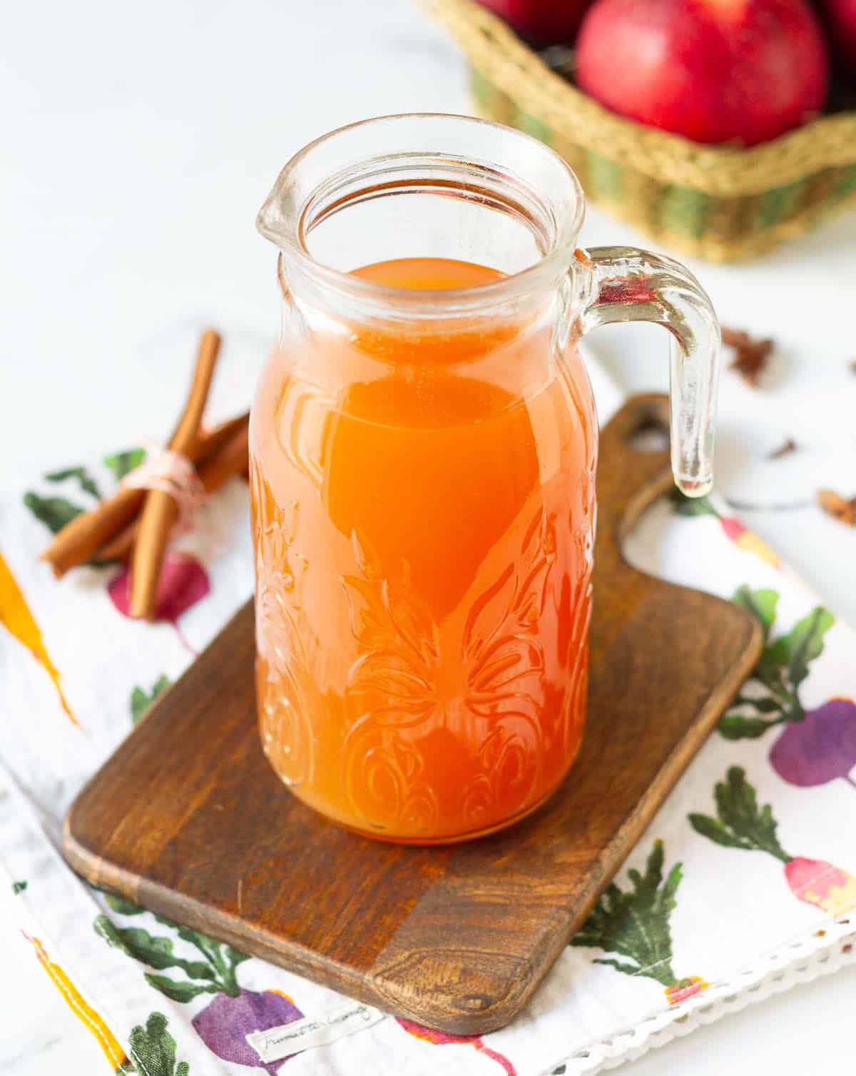 Apple cider in a clear glass pitcher on a wooden board.