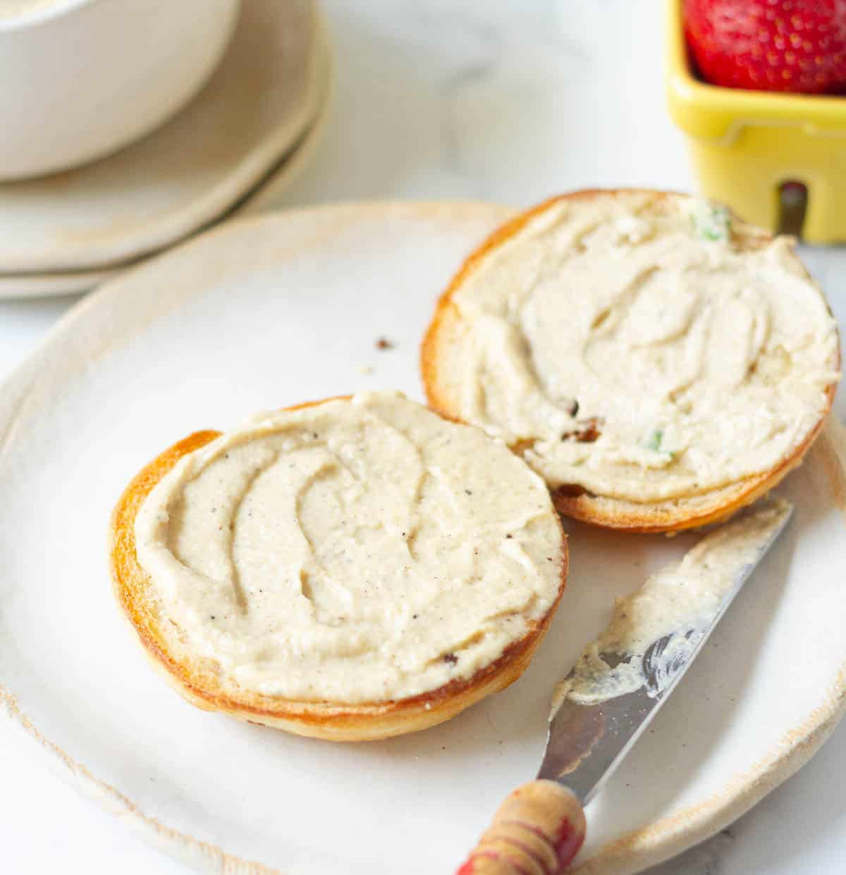 cashew cheese smeared over a sliced bagel on a plate with a knife.