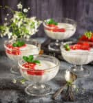 4 bowls of panna cotta with strawberry sauce on top and flowers and spoons around.