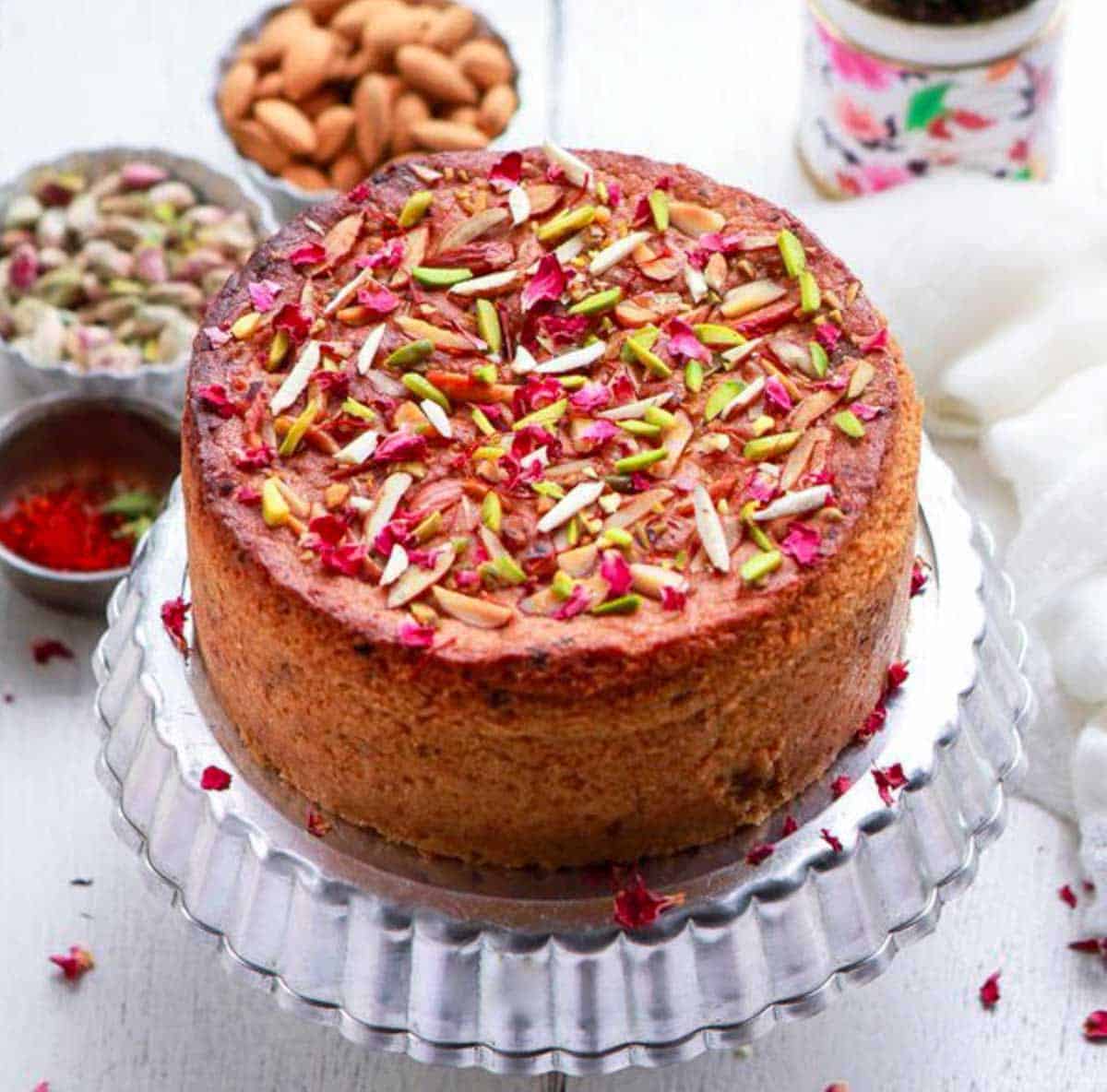 Cake with nuts and rose petals on a cake stand with ingredients in the background.