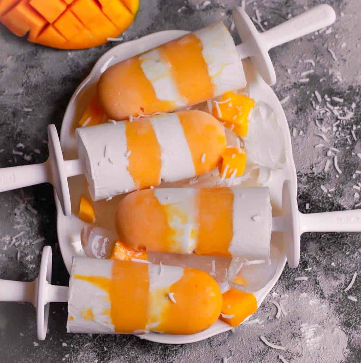 Mango coconut popsicles on a tray with a cut mango on the side.