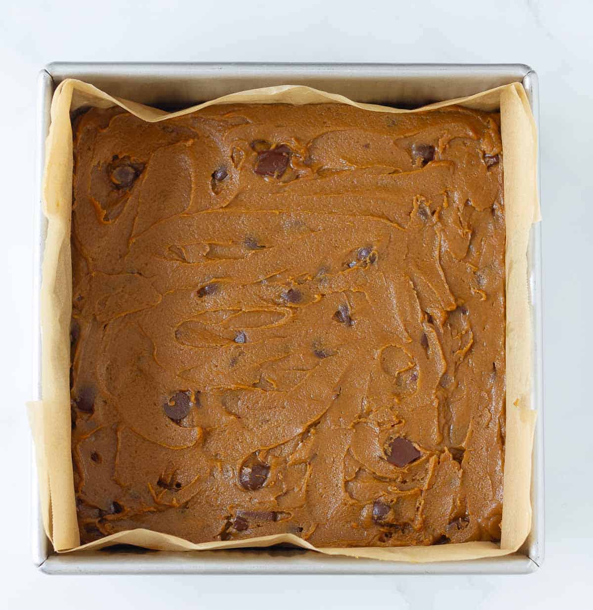 Batter with chocolate chips in a parchment paper lined square baking tin.