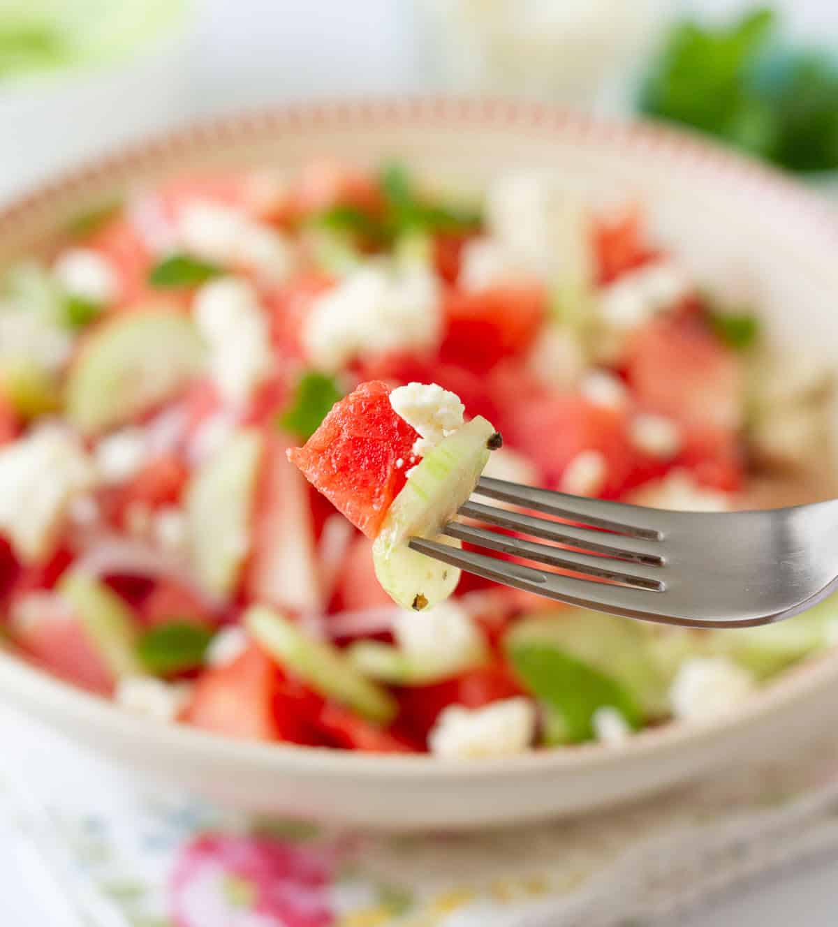 A fork with cucumber, watermelon and feta cheese with a salad bowl in the background.