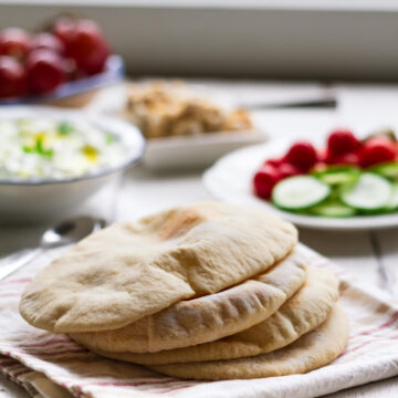 Pita Breads stacked on a kitchen towel with dips and salads in the background.