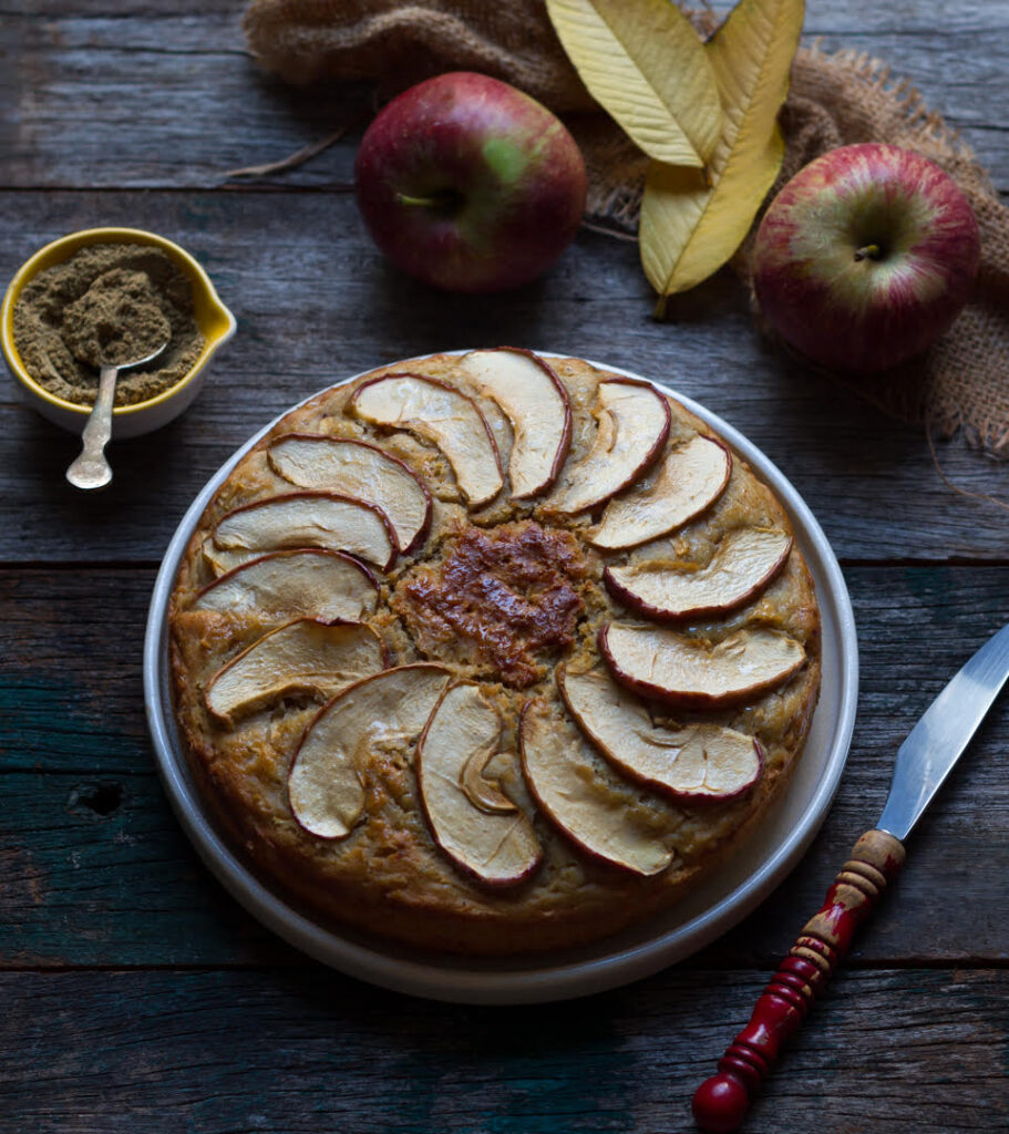 Baked apple cake on a serving plate with a knife and chai masala in the background.