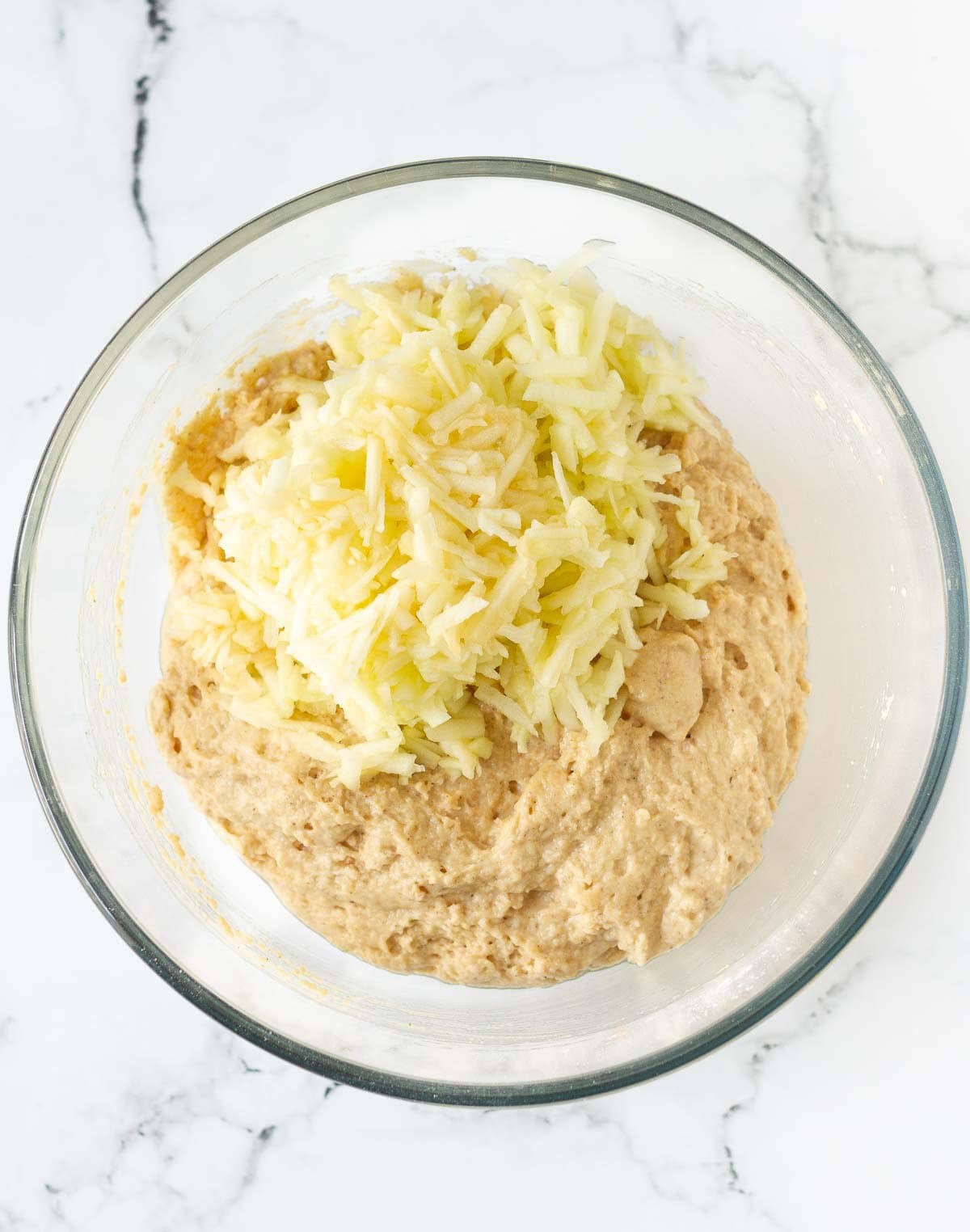 Grated apples on top of the cake batter in a glass bowl.