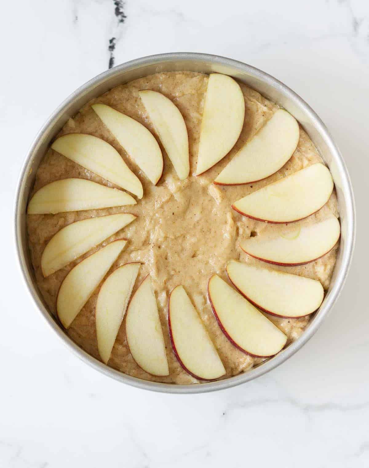 apple cake batter in a round cake tin topped with apple slices.