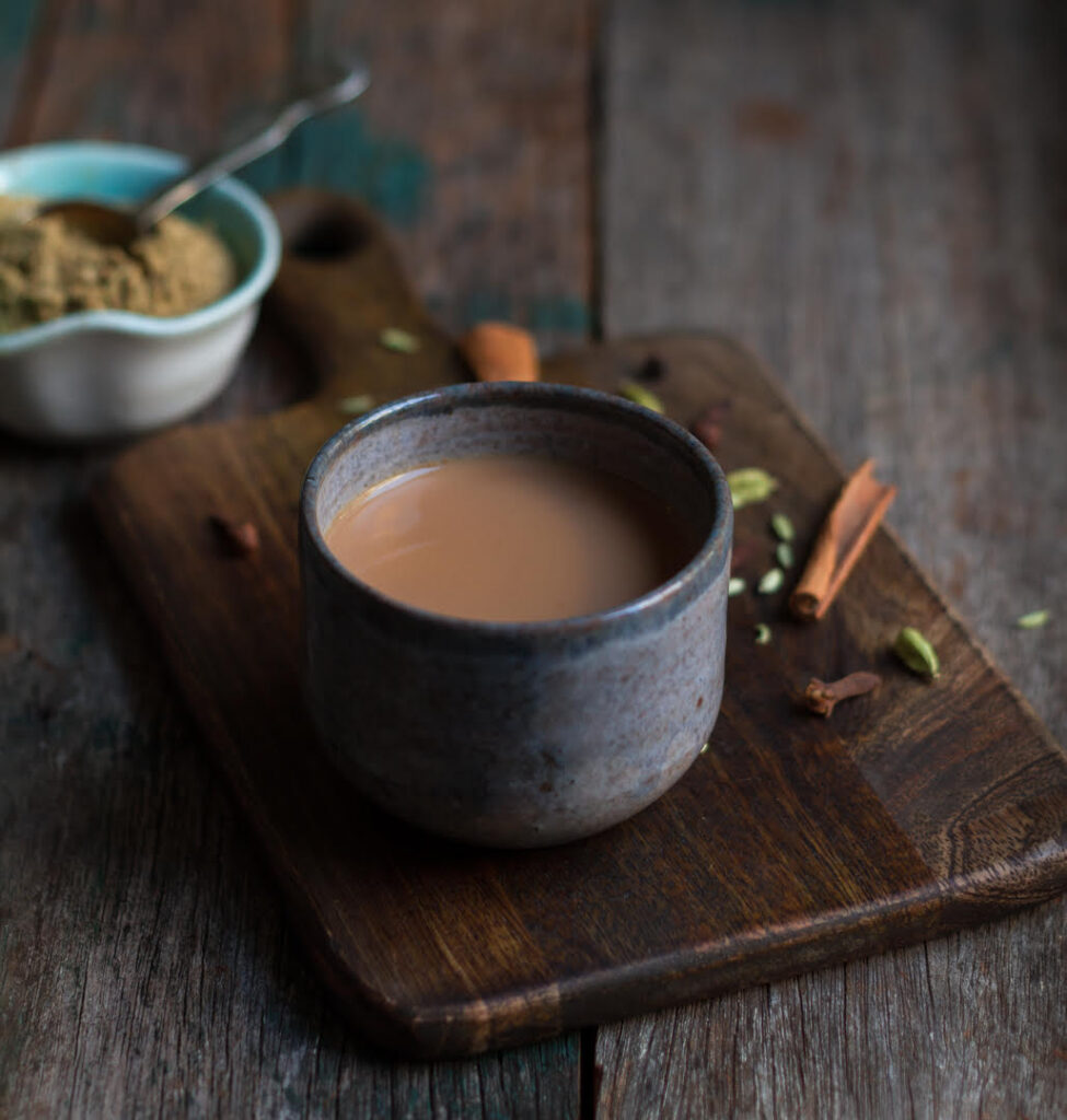 A cup of masala chai or tea on a wooden board with ingredients scattered.