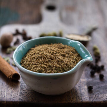 chai masala in a small bowl on a wooden board with ingredients in the background.