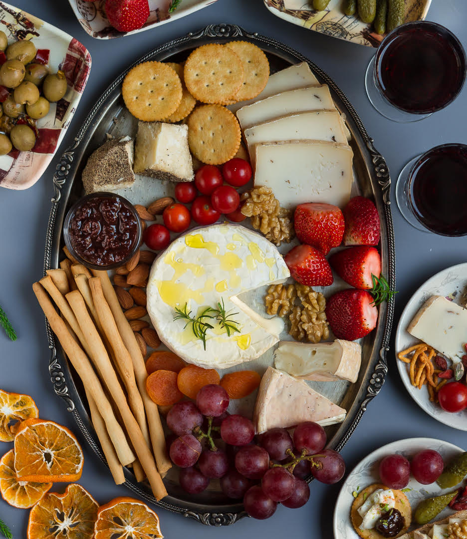 A silver cheese platter with cheese, soup sticks, crackers,fruits, olives, nuts and jam.