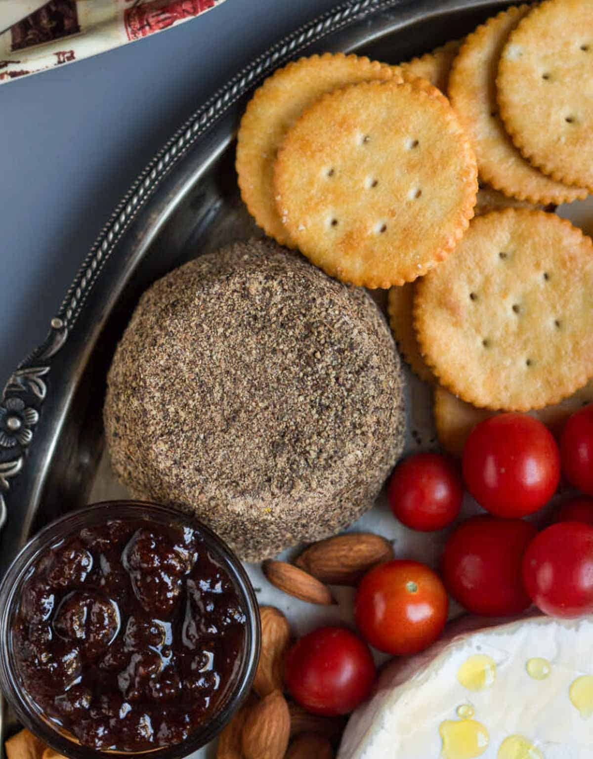 Garlicky pepper-crusted cheese with crackers, cherry tomatoes, jam on a silver platter.