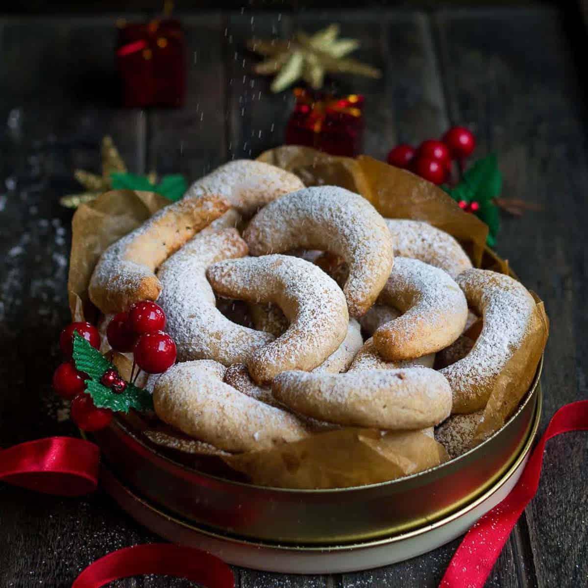 Crescent shaped cookies dusted with sugar in a round cookie box with christmas decor around.
