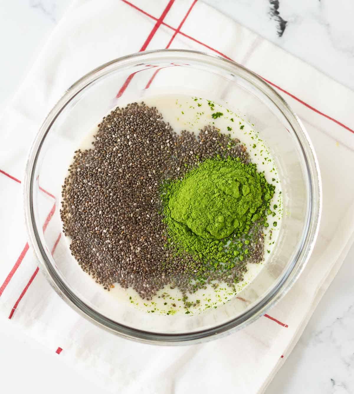 Chia seeds, matcha powder, maple syrup, vanilla extract and almond milk in a glass bowl.