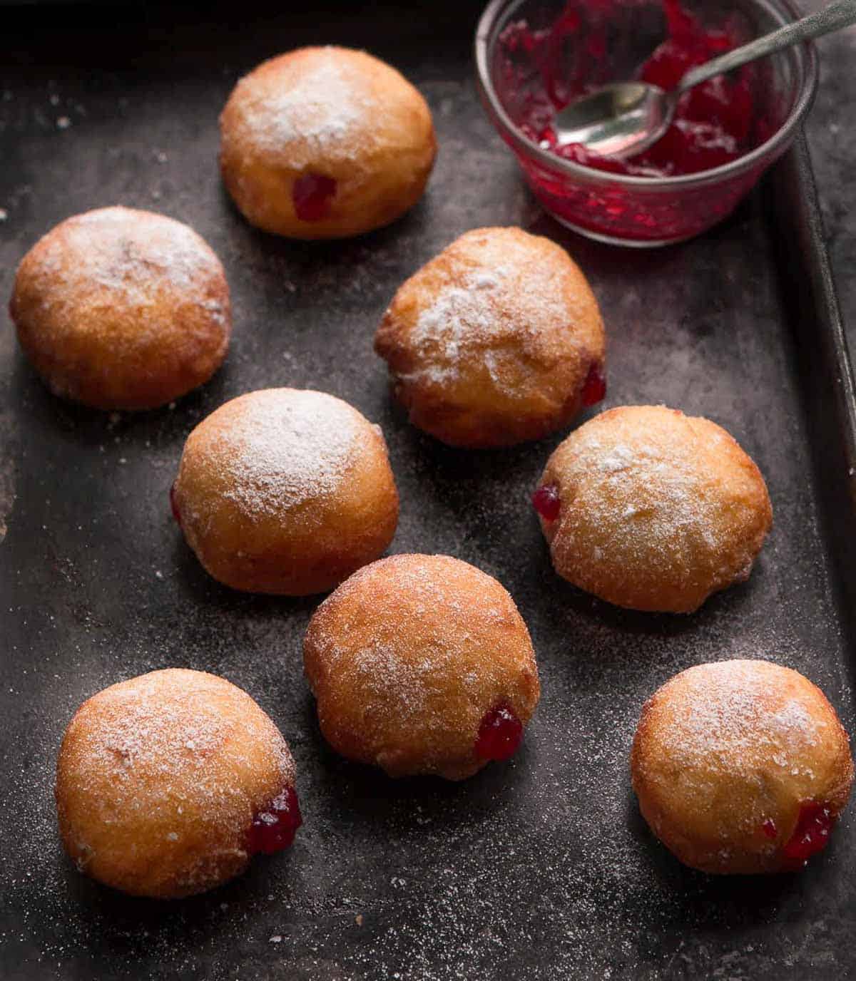 strawberry jam filled donuts on a tray with a bowl of strawberry jam on the side.