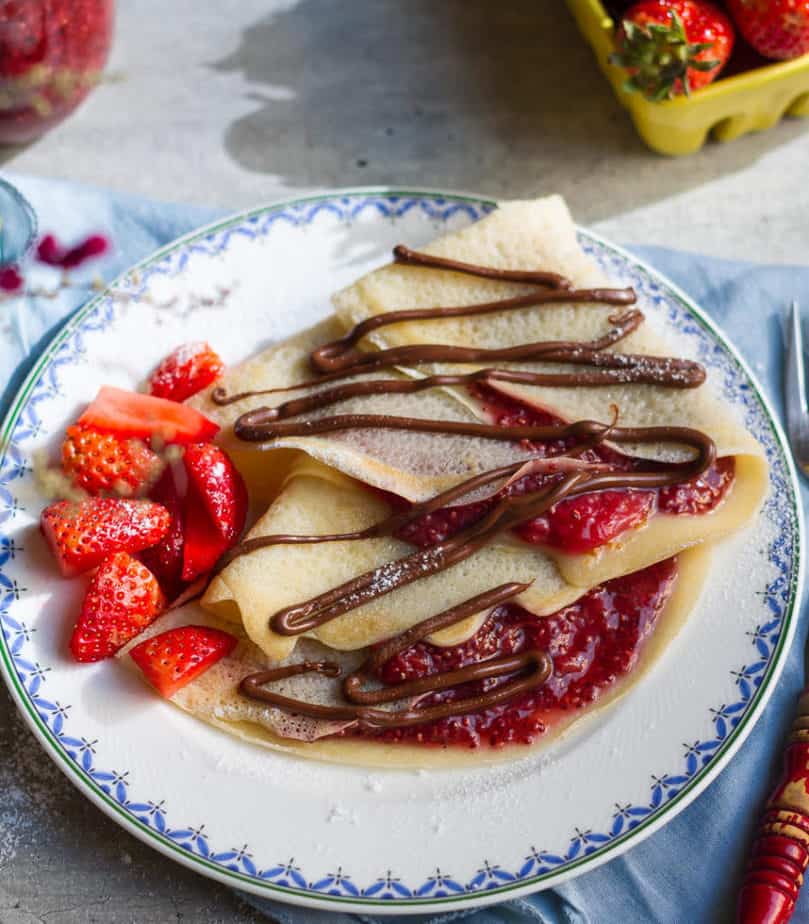 Crepes filled with strawberry jam and drizzled with Nutella on a plate with fresh strawberries.