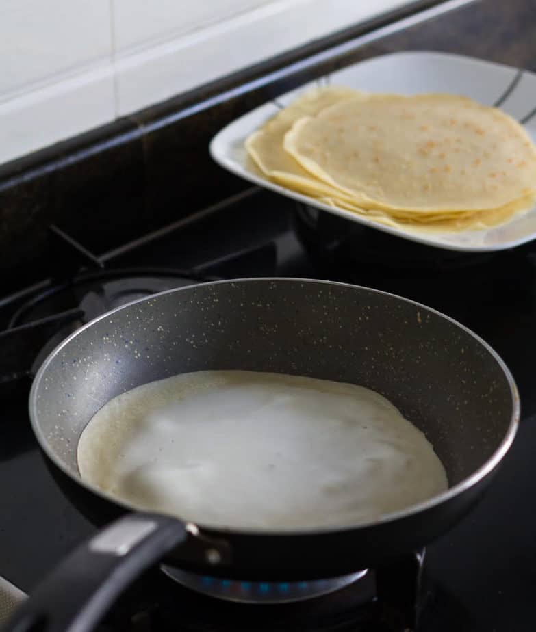 cooking a crepe in a non stick skillet with crepes on a plate in the background.