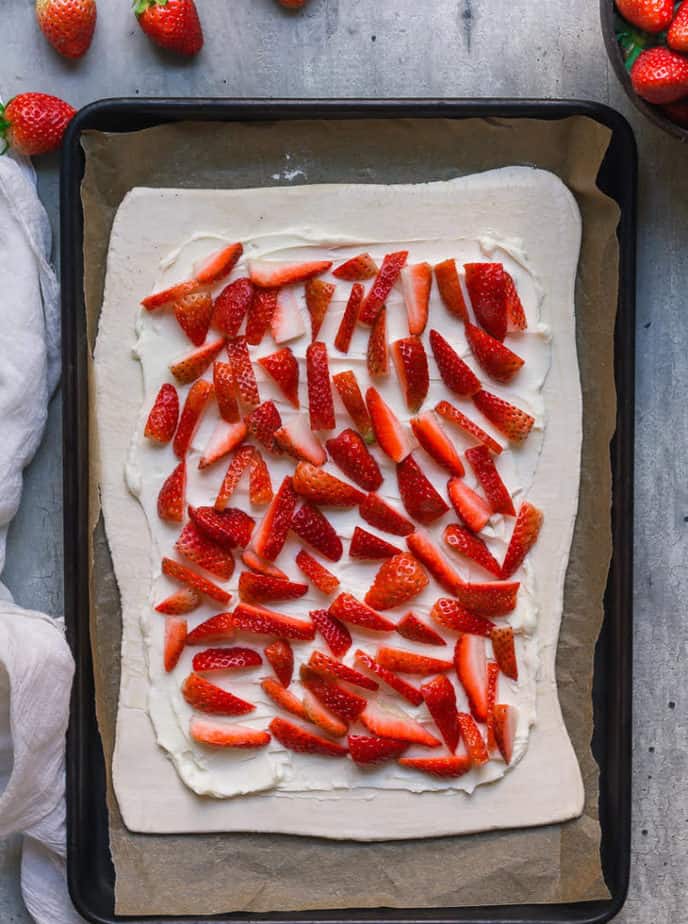Puff pastry topped with cream cheese and strawberries on a parchment-lined baking tray.