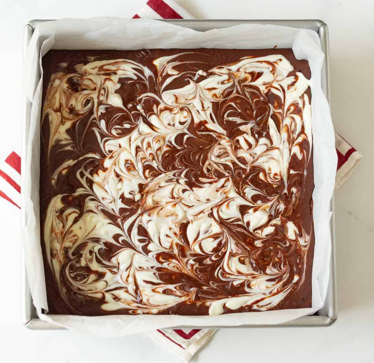Swirled brownie and cheesecake batters in a square pan lined with parchment paper.