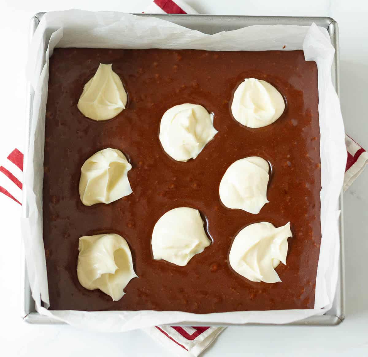 Spoonfuls of cheesecake mixture on top of brownie batter in a square baking pan.