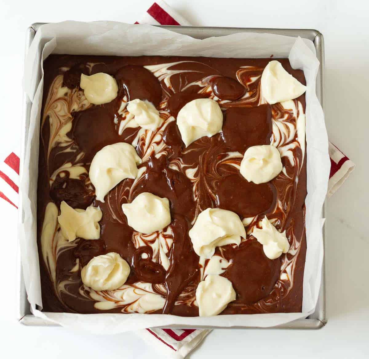 Cheesecake and brownie batter swirls in a square baking pan lined with parchment paper.