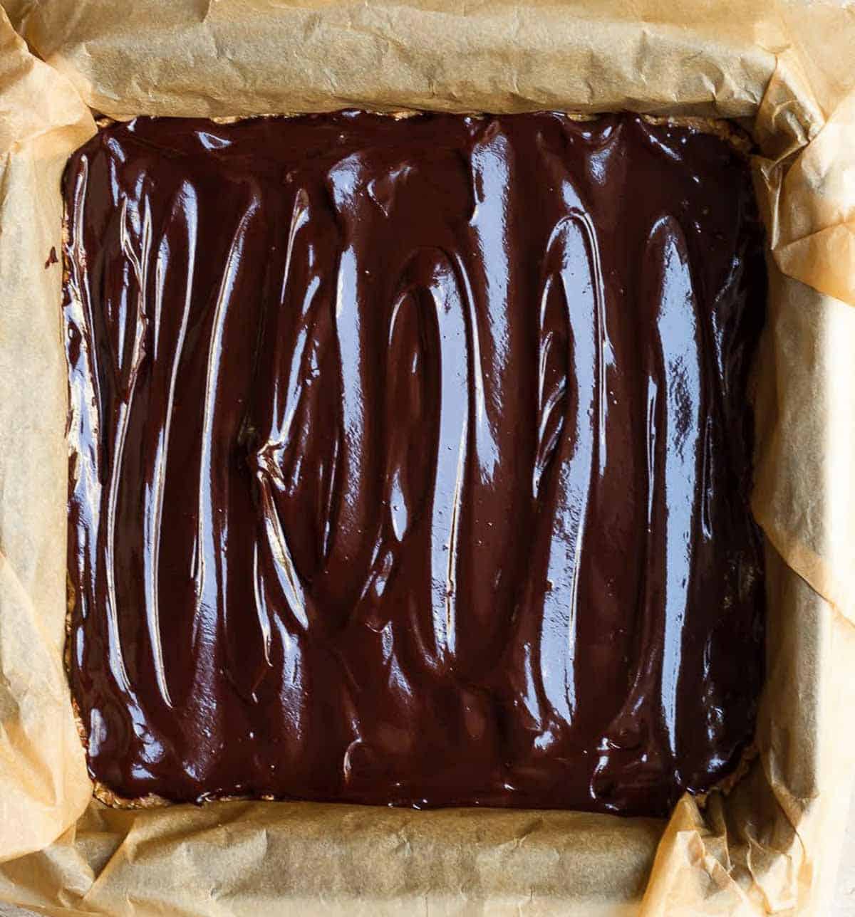Layer of melted chocolate on the oats base on parchment paper in a square pan.