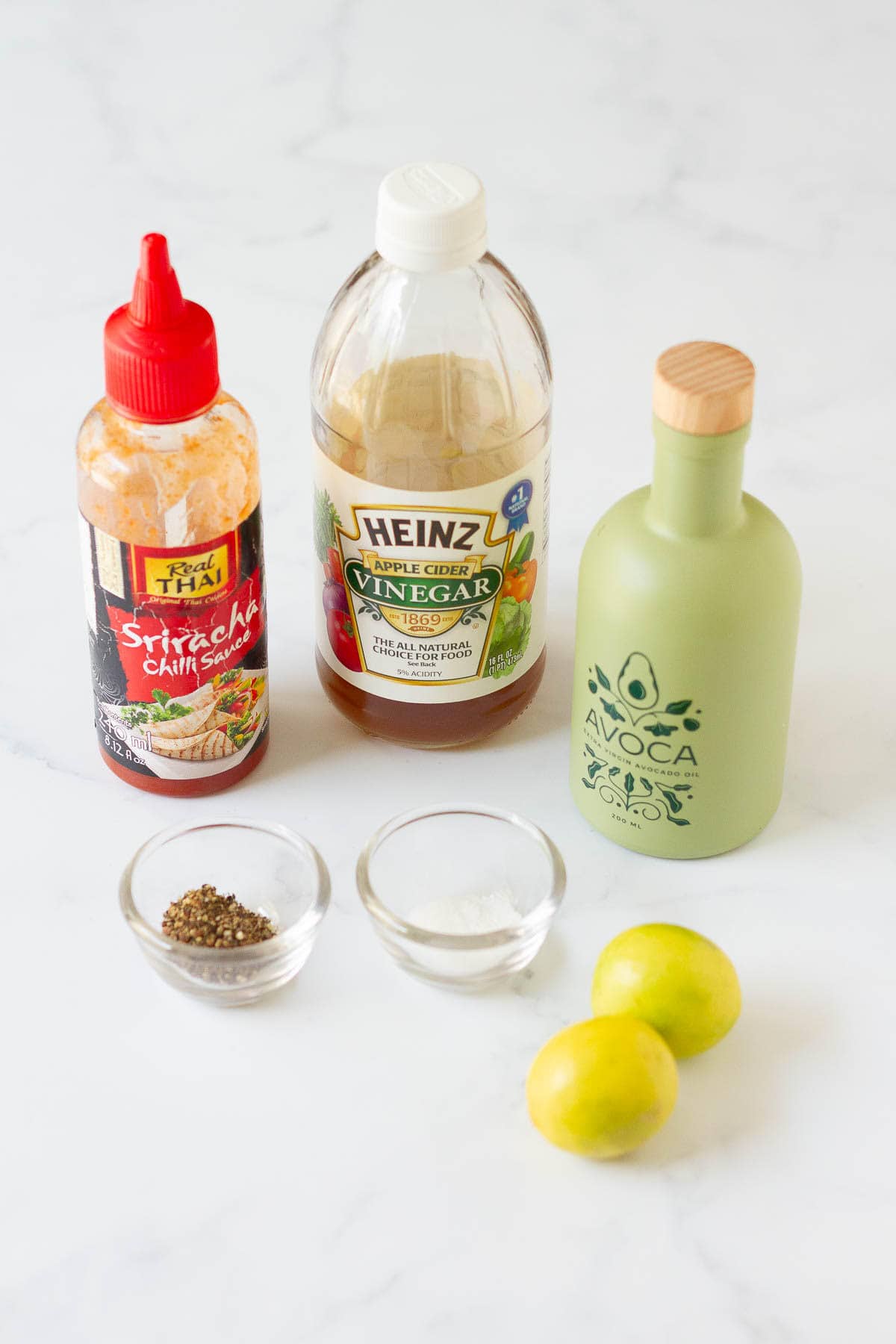 Sriracha, avocado oil, vinegar, lemons, salt , and pepper on a white surface.
