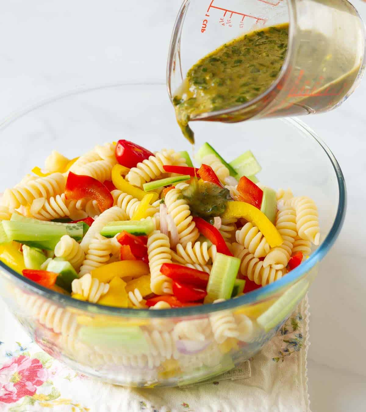 Salad dressing getting poured over veggies and pasta in a bowl.