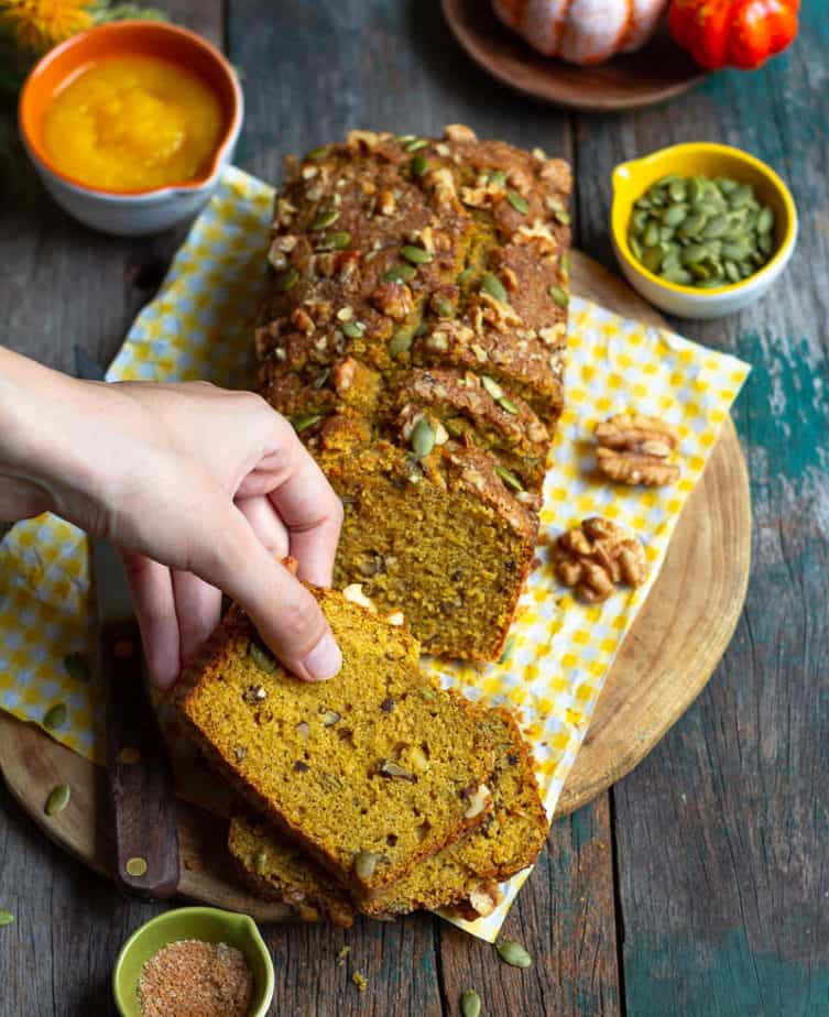 A hand picking up a slice of pumpkin bread on a wooden board with a knife.