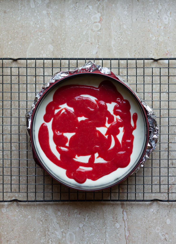 Strawberry sauce on top of the cheesecake batter in a round cake pan.
