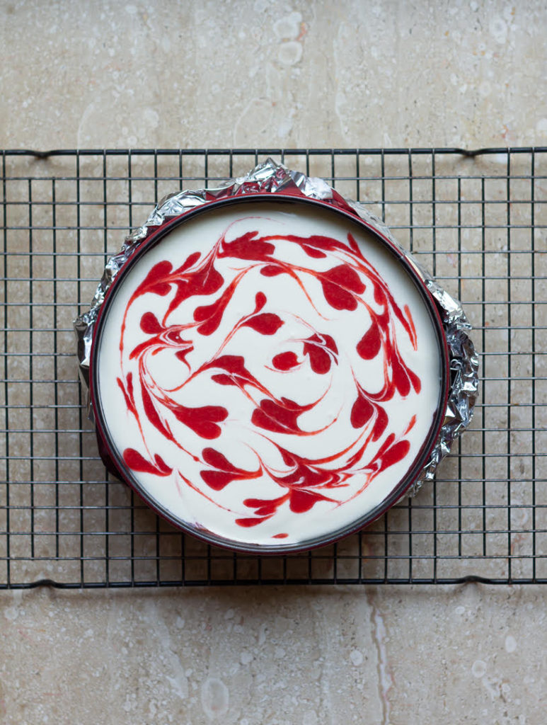 Strawberry sauce swirls on top of the cheesecake batter in a cake pan.