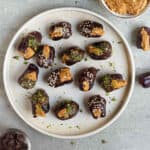 Peanut butter stuffed dates dipped in chocolate and topped with pistachios on a plate.
